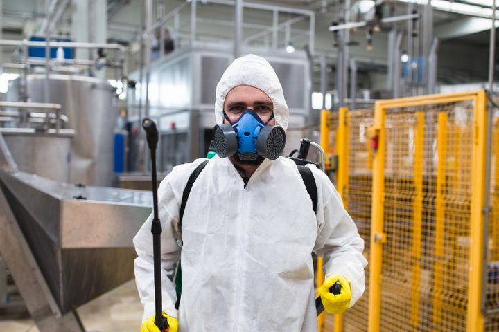 Técnico aplicador con buzo, máscara de filtros y lanza de aplicación, de pie en el pasillo de una planta industrial