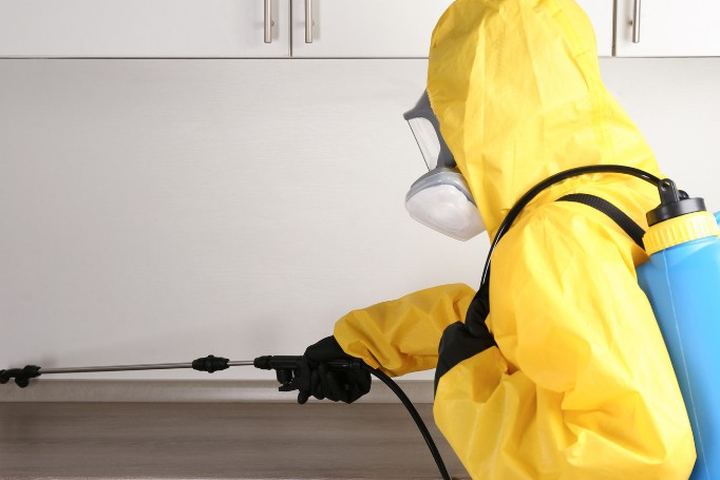Aplicador con buzo amarillo y máscara de filtros trata con lanza larga el hueco bajo los muebles de una cocina en un restaurante