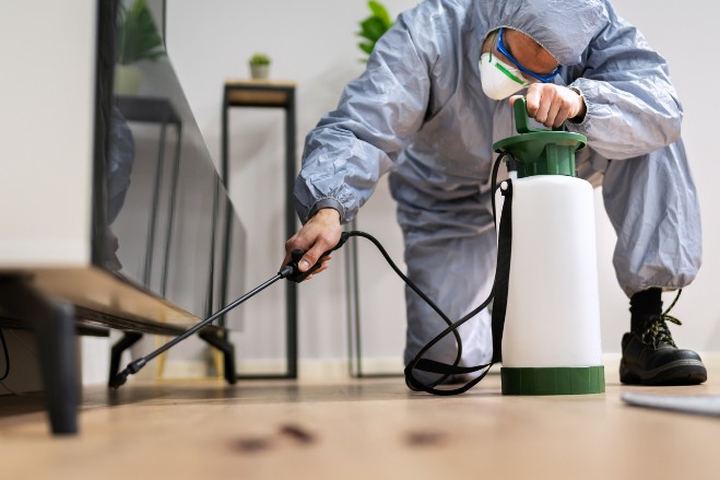 Técnico con buzo gris y mascarilla aplica con lanza bajo el mueble de la televisión de un salón