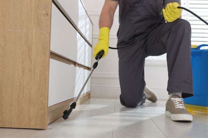 Aplicador arrodillado con guantes de nitrilo trata con lanza el zócalo inferior de un mueble de cocina de madera clara