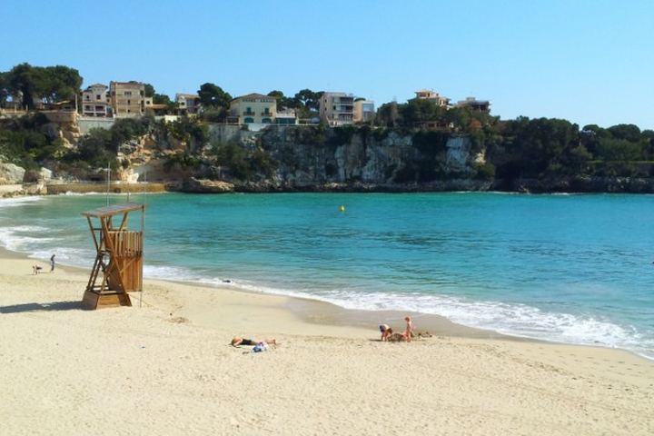 Cala de arena con torre de vigilancia de madera, agua turquesa y chalets sobre el acantilado del litoral de Llevant