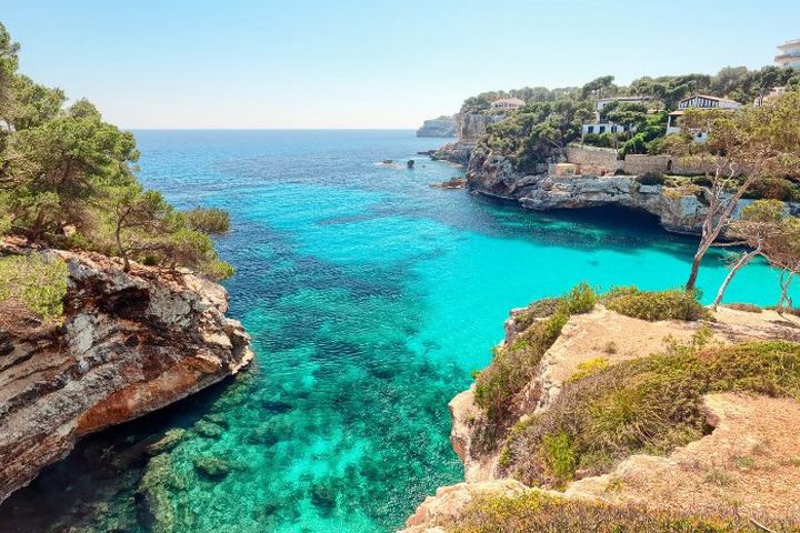Cala de aguas turquesas encajada entre acantilados de marés, con pinar y casas de veraneo en la parte alta